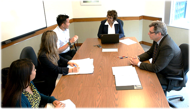 A man gestures as he testifies at a boardroom table. The adjudicator takes notes as she listens. Beside the man sits his legal representative and his witness. Across the table sits the respondent.