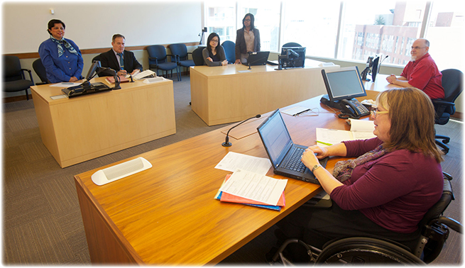 Une arbitre est assise à un bureau, face à l'appelante et à l'intimée et à leurs représentants légaux. La représentante légale de l'appelante et l'intimée sont debout derrière leur bureau. Un homme est assis à un bureau à droite de l'arbitre.