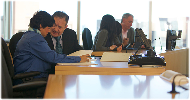 Un homme et une femme sont assis dans une salle d'audience et examinent leurs documents. À l'arrière-plan, d'autres personnes sont assises à leur bureau.