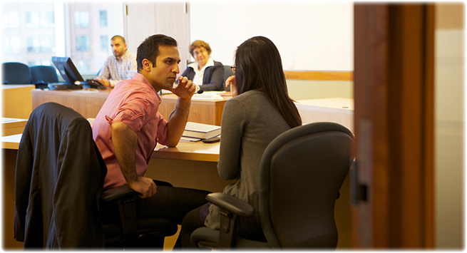 A man and woman talk to each other at their desk. In the background, two adjudicators observe them.