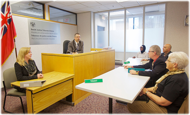 A member sits at an elevated desk in a hearing room. Across from him sit the landlord and their legal representative at one desk, and the tenant and their legal representative at another desk. Beside the member sits a woman at desk.