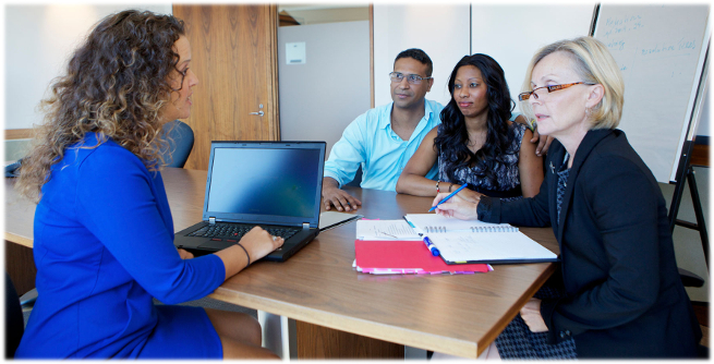 At a boardroom table. A lawyer with a laptop speaks to an adjudicator at the head of the table. On the other side of the table, a couple who are the applicants, watch.