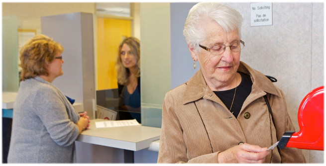 A woman takes a number from a machine. In the background, another woman receives service at the desk.