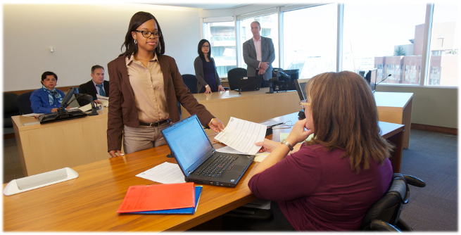 A woman passes a document to an adjudicator at a dias. In the background, other people are sitting at desks.